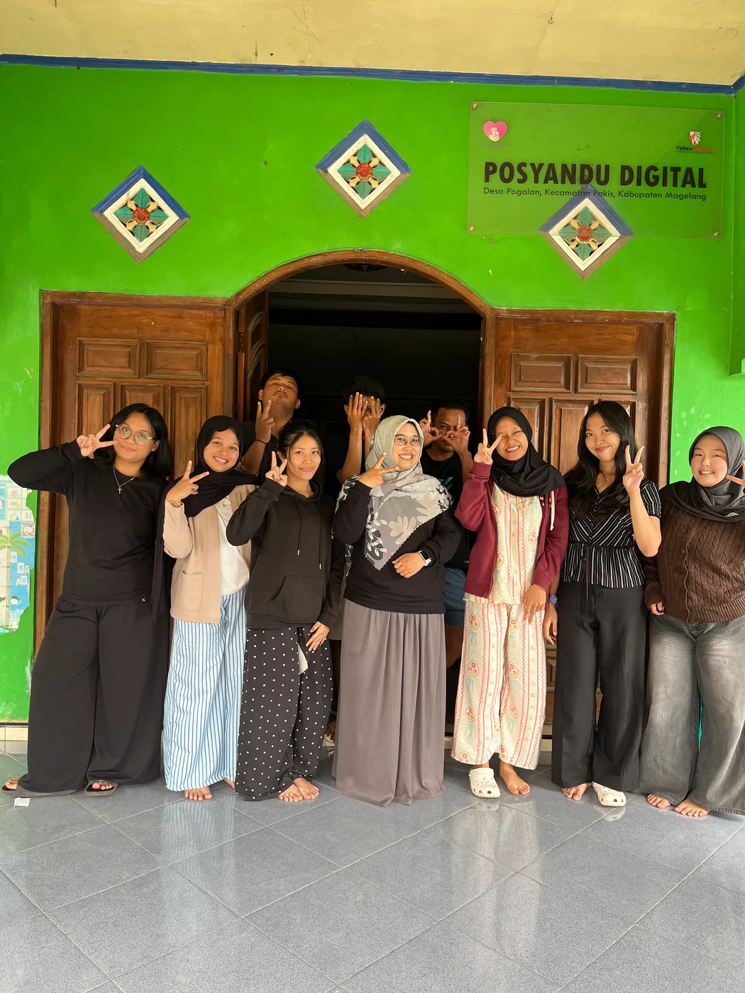 Anggota kelompok berfoto santai bersama di depan pintu posko berwarna hijau